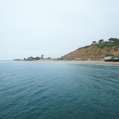 Coastal scene with calm water, a sandy beach, and a hillside with trees and buildings under a cloudy sky.