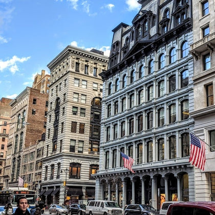 Urban street with historic multi-story buildings, American flags, cars parked, and pedestrians under a blue sky.