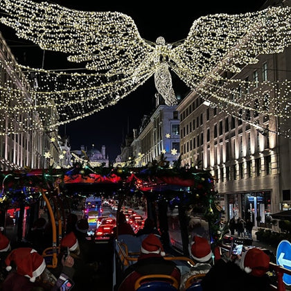 A festive street at night with illuminated angel decorations, viewed from a bus with passengers wearing Santa hats.