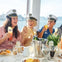 Three people in sailor hats enjoy drinks and laughter at a table with food and champagne, with a scenic water view behind.