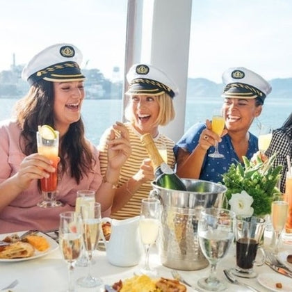 Three people in sailor hats enjoy drinks and laughter at a table with food and champagne, with a scenic water view behind.