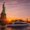 A sightseeing boat labeled 'NY Skyline' passes by the Statue of Liberty at sunset, with a cityscape visible in the background.