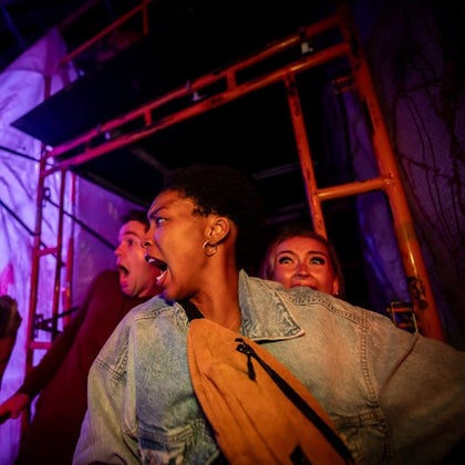 Three people in various expressions of surprise or fear, with scaffolding and dim lighting in the background.