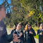 A group of people outdoors in a forested area, one person speaking while others listen. Sunlit trees are in the background.