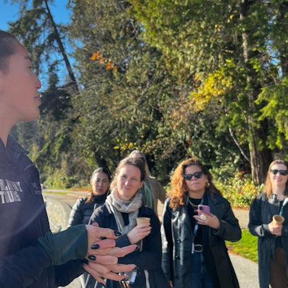 A group of people outdoors in a forested area, one person speaking while others listen. Sunlit trees are in the background.