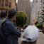 Urban street scene with a group of people in the foreground and tall buildings, traffic, and a tree in the background.