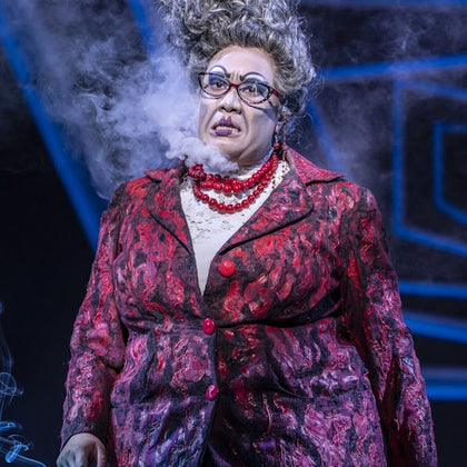 A person in theatrical makeup and costume, wearing a patterned suit and beads, with dramatic lighting and smoke effects.