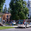 A tour tram with seated passengers passes through a town-like area with trees and buildings, including a church.