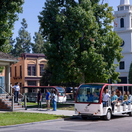 A tour tram with seated passengers passes through a town-like area with trees and buildings, including a church.