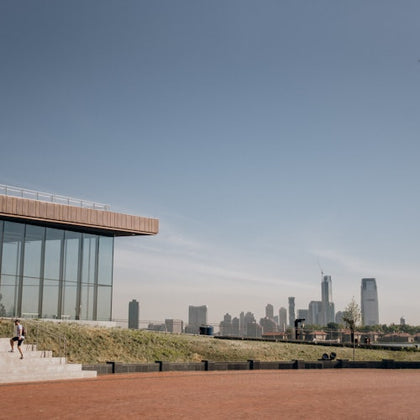 Modern building with glass facade on the left, steps and a person walking down. City skyline visible in the background.
