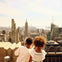 Two children stand on a rooftop observation deck, looking at skyscrapers through a coin-operated viewer on a sunny day.
