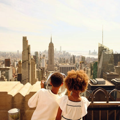 Two children stand on a rooftop observation deck, looking at skyscrapers through a coin-operated viewer on a sunny day.