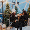 Two people in winter attire toast with drinks while seated inside a festive, snow-covered dome decorated with Christmas trees.