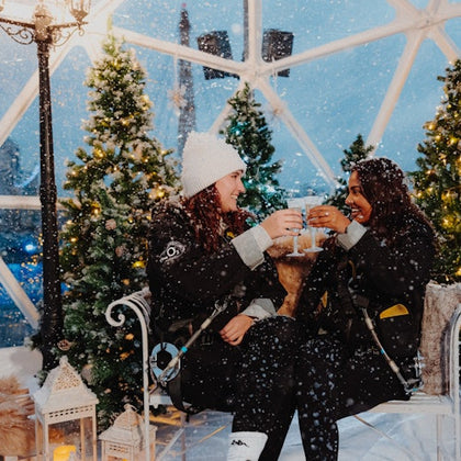 Two people in winter attire toast with drinks while seated inside a festive, snow-covered dome decorated with Christmas trees.