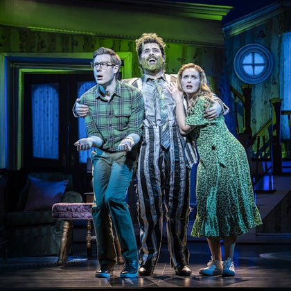 Three actors in a theatrical performance stand on a stage, with dramatic lighting and a detailed interior set as a backdrop.