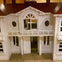 A miniature white wooden house with a red roof, circular windows, and a small balcony on a tiled floor indoors.