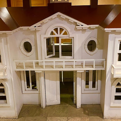 A miniature white wooden house with a red roof, circular windows, and a small balcony on a tiled floor indoors.