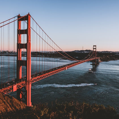 Golden Gate Bridge extending over water with boats below, against a backdrop of hills and a clear sky.