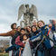 A group of people in safety harnesses take a selfie near a stone statue with wings, outdoors with a city view in the background.