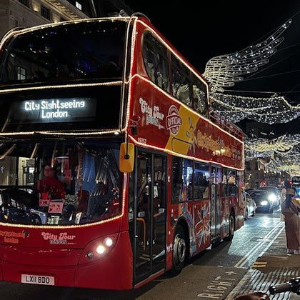 A red City Sightseeing London double-decker bus drives through a night-lit street with festive lights overhead.