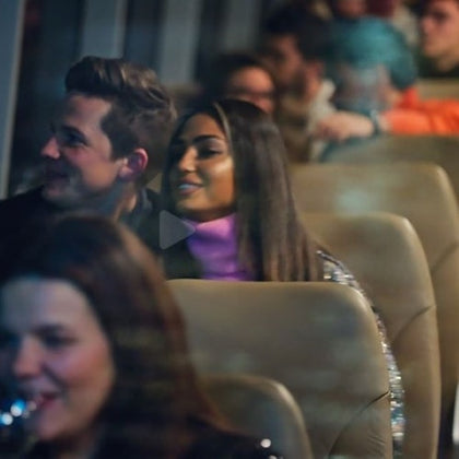 People sitting on a bus in soft lighting, smiling and conversing. The focus is on a man and a woman in the middle of the image.