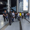 A group of people standing on a modern building walkway near large windows and structural columns, with a plane in the distance.