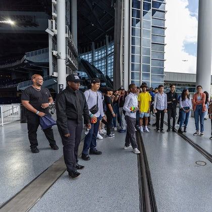 A group of people standing on a modern building walkway near large windows and structural columns, with a plane in the distance.