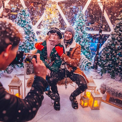 A couple poses for a photo in a snowy holiday setting, surrounded by Christmas trees and glowing lanterns, as another person takes their picture.