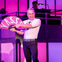 A man points to a pink fan that reads "YASSS" while performing on stage with a lit purple and pink background, musicians behind him.