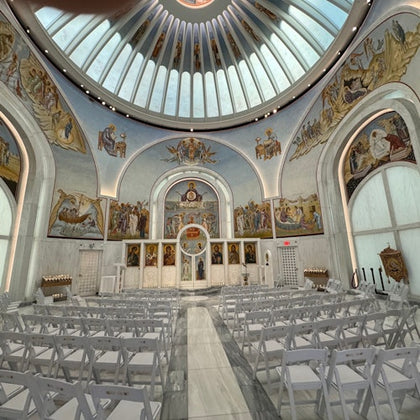A church interior with a domed ceiling, religious murals, white chairs, and an ornate altar at the front.