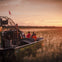 People on an airboat ride through a marsh at sunset, with a person standing in the front and others seated, viewing the landscape.