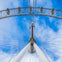 Upward view of a Ferris wheel structure against a cloudy blue sky, showing the wheel spokes and passenger capsules.