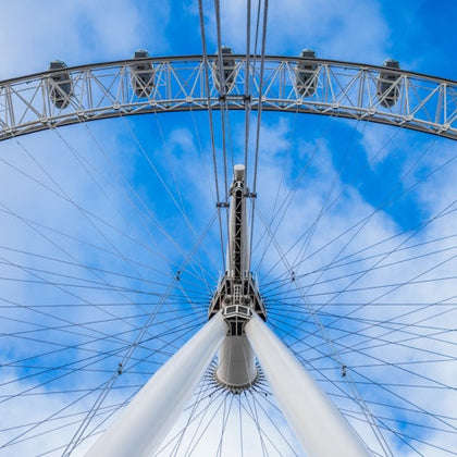 Upward view of a Ferris wheel structure against a cloudy blue sky, showing the wheel spokes and passenger capsules.
