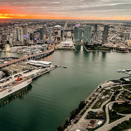 Aerial view of a cityscape at sunset with high-rise buildings, a bridge, a docked cruise ship, and a waterfront area.