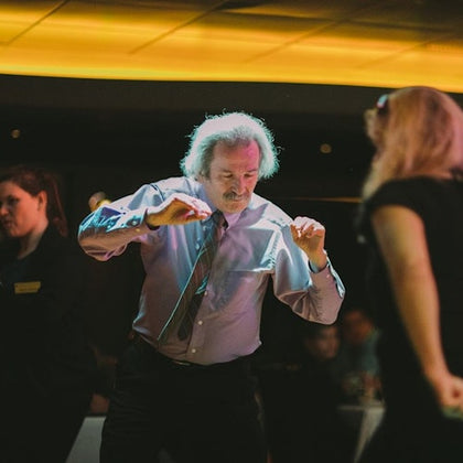 A man in a tie dances energetically with two other people in a dimly lit room with a yellow-lit ceiling.