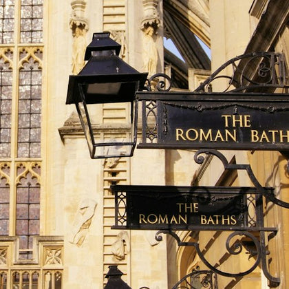 Signs reading "The Roman Baths" hanging near an ornate building facade with stained glass and sculptures.