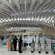 A modern transportation terminal with curved ceiling beams, police officers, and turnstiles in the foreground, people in the background.