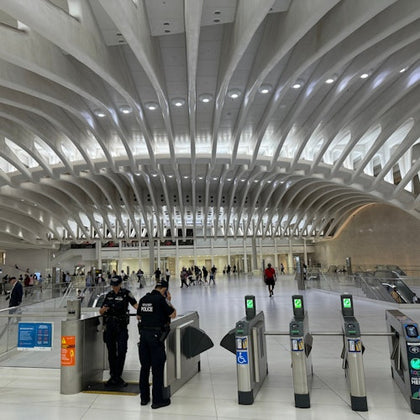 A modern transportation terminal with curved ceiling beams, police officers, and turnstiles in the foreground, people in the background.