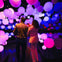 Two people walk through a colorful installation of glowing spheres in a dark room with vivid purple and pink lighting.
