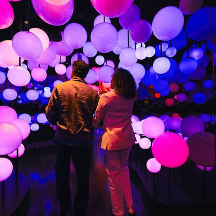 Two people walk through a colorful installation of glowing spheres in a dark room with vivid purple and pink lighting.