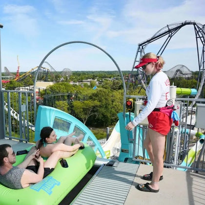 Two people sit in a green inflatable raft at the top of a water slide, with a lifeguard standing nearby. Roller coasters are visible in the background.