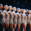 A lineup of female dancers in sparkly, silver fringe dresses, performing on stage under warm lights.
