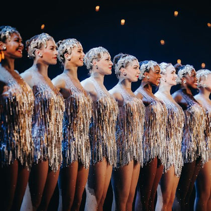 A lineup of female dancers in sparkly, silver fringe dresses, performing on stage under warm lights.
