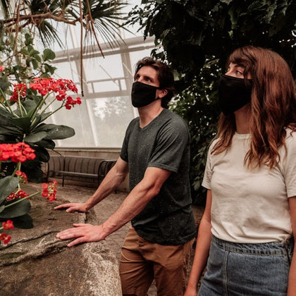 Two people wearing black face masks observe red flowers inside a greenhouse, hands resting on a tall stone ledge.