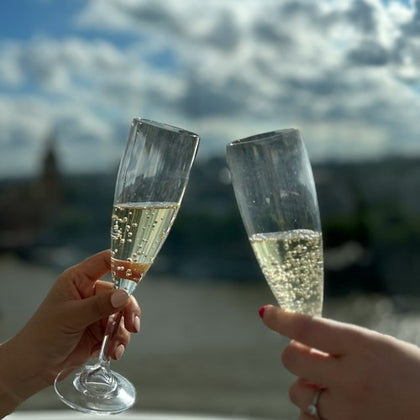 Two hands clinking champagne glasses against a blurred outdoor backdrop under a partly cloudy sky.