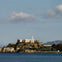 Island with a large building and lighthouse, surrounded by water and distant hills under a partly cloudy sky.