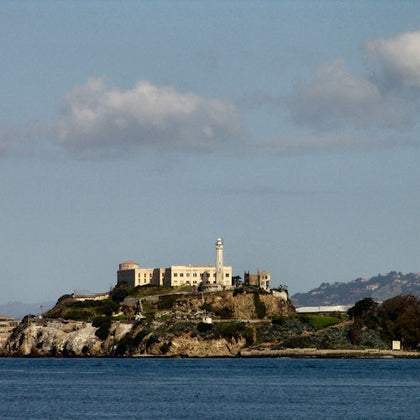 Island with a large building and lighthouse, surrounded by water and distant hills under a partly cloudy sky.