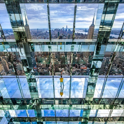 A person stands on a glass floor in a mirrored observation space, with a cityscape and the Empire State Building visible.