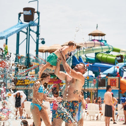 People enjoying a water park with slides and water play areas, featuring a person lifting a child in the air.