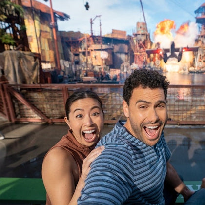 A man and a woman excitedly pose in front of a theme park stunt show with explosions and water effects in the background.